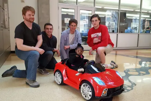 Students around A “Go-Baby-Go” car with a young child sitting in the car. 