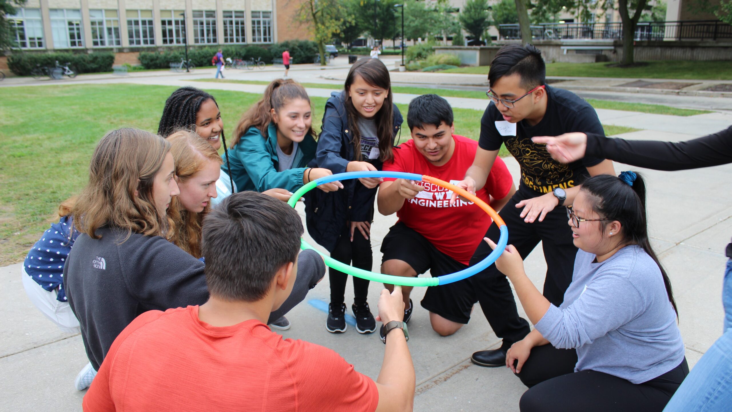 Nine students outside crouching down hold up a hula hoop with one finger each.