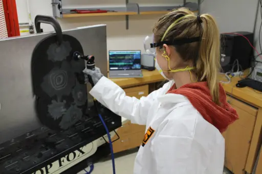 Woman with ponytail with her back to the camera, wearing white labcoat, mask and lab goggles,working in a lab grinding a vertical surface as she looks at a computer screen in the distance.