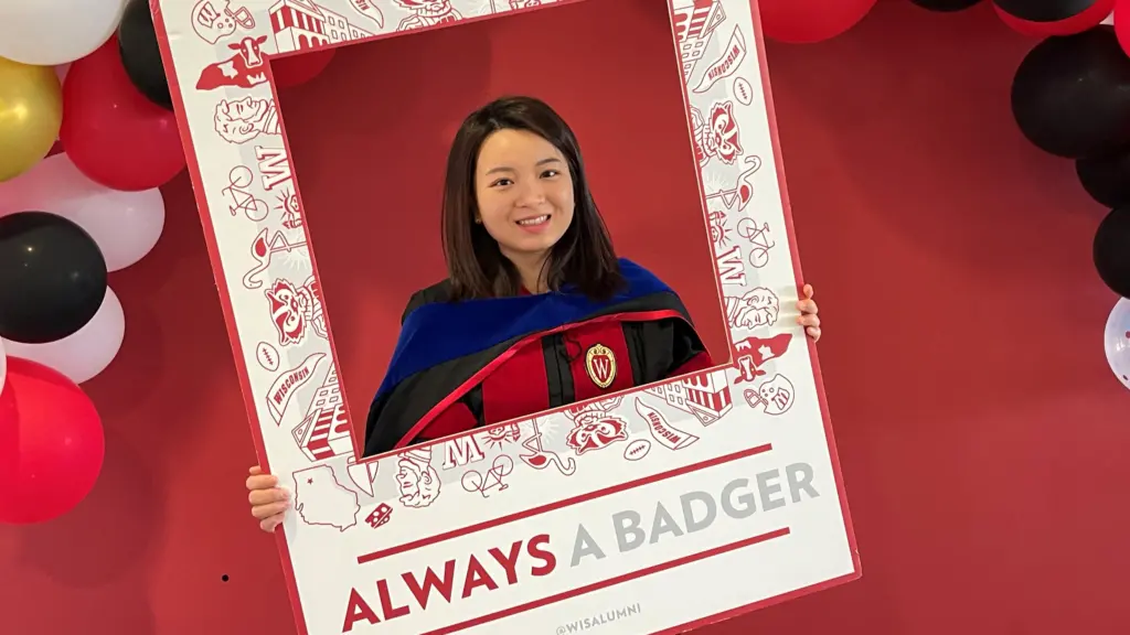 A PhD graduate dressed in a doctoral gown holds up a frame that reads "always a Badger" during a celebration hosted by the UW-Madison Civil and Environmental Engineering Department in spring 2023.