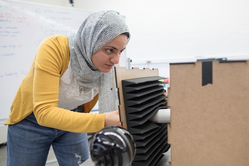 ECE Grad Student in yellow sweater and grey patterned head scarf closes end of black, foam-covered, anechoic chamber that contains a white PVC tube and sits next to a black ball microphone