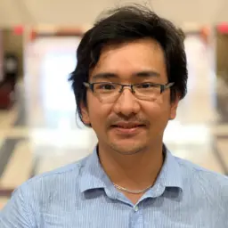 head shot of a man wearing glasses and a button-up shirt, with a blurry building atrium in the background