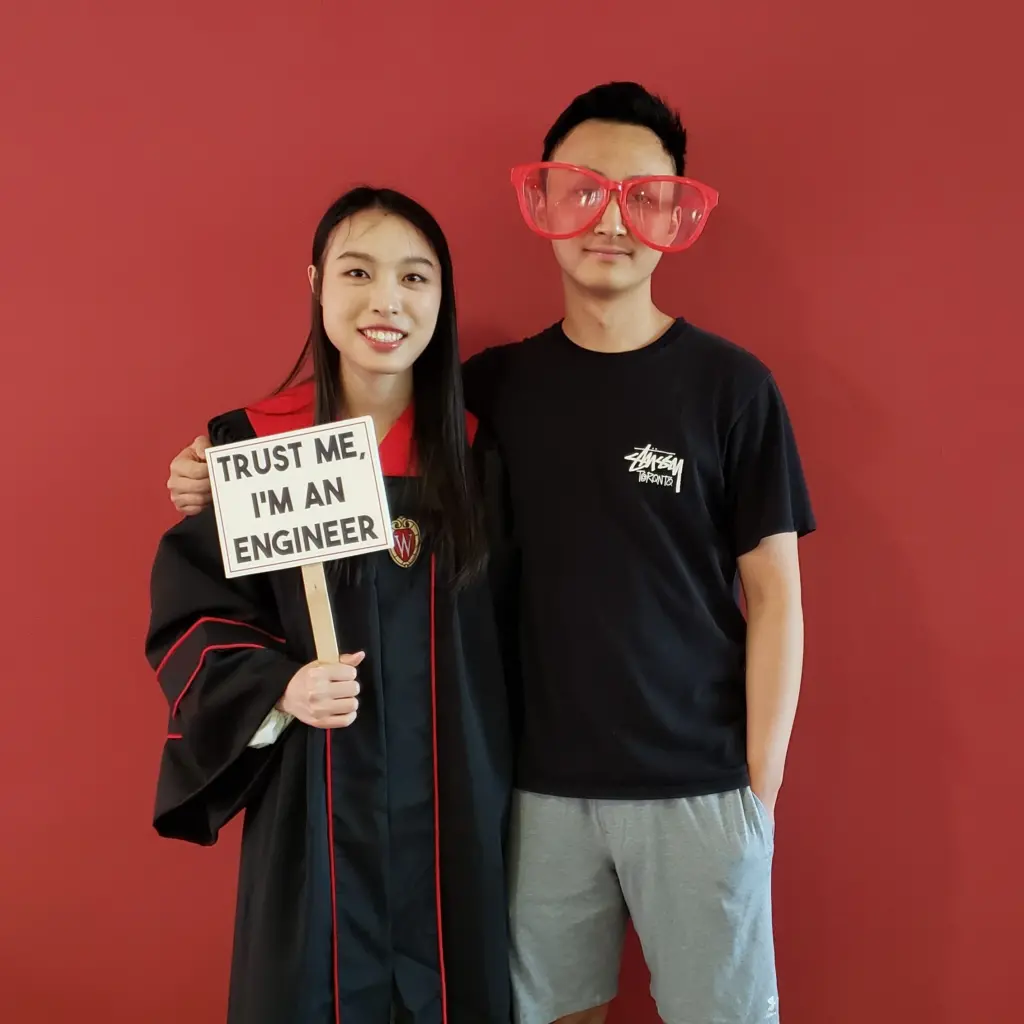 2022 Graduate in her commencement gown stands with friend holding a sign that reads, "Trust me, I'm an Engineer"