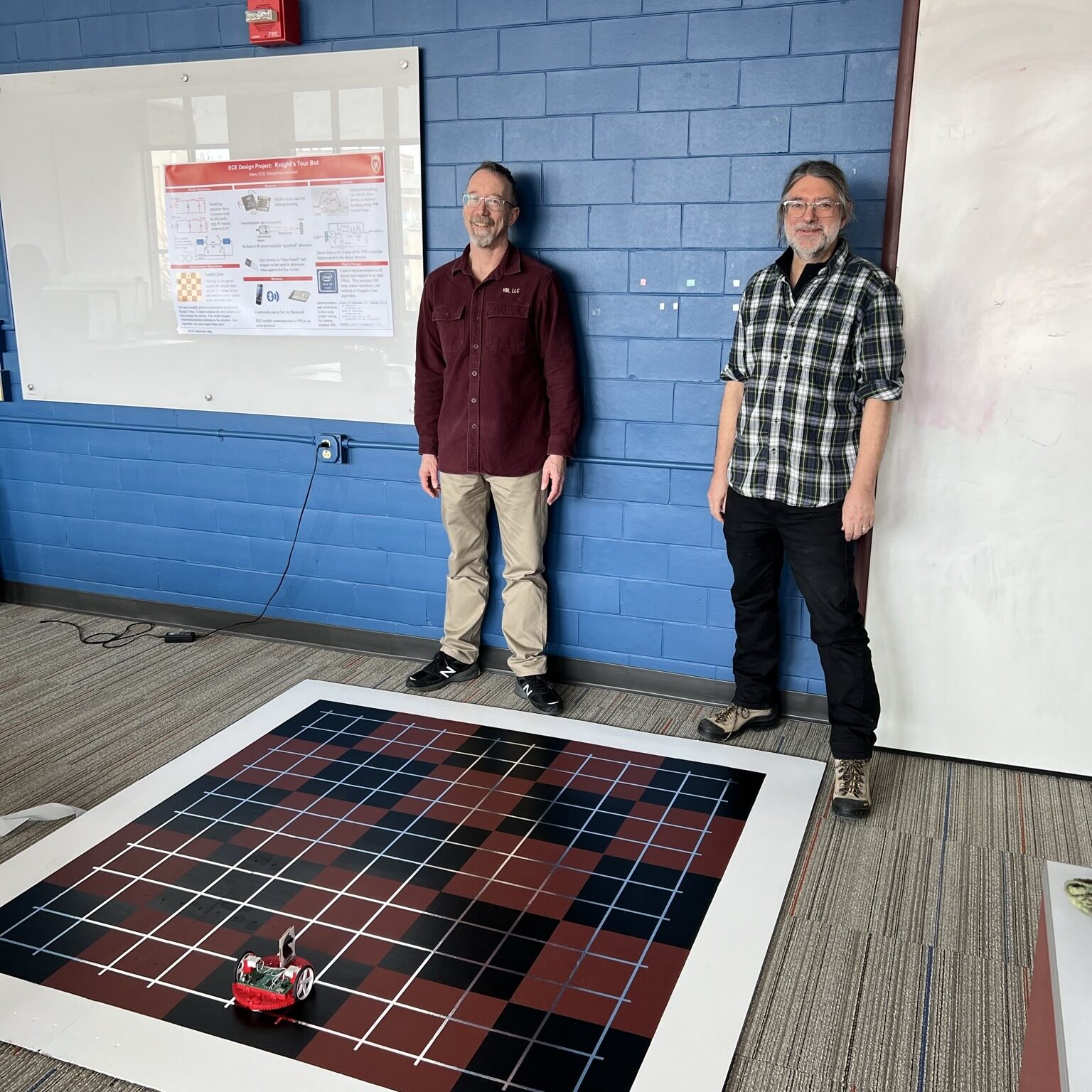 Eric Hoffman and Bill Sethares stand behind a chess board on the floor with small wheeled object on the board