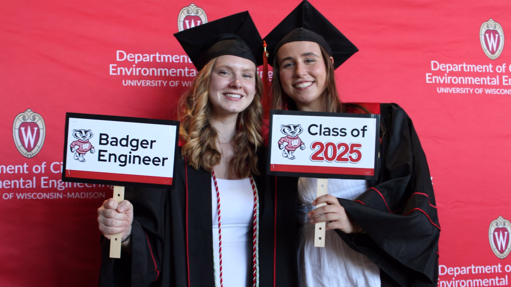Two graduates wearing graduation caps and gowns pose with photo props.