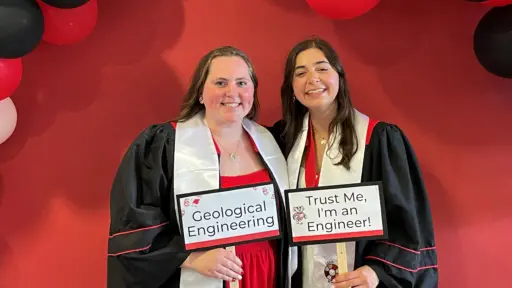 Two bachelors degree graduates from the UW-Madison Department of Civil and Environmental Engineering hold up signs that read "Geological Engineering" and "Trust Me, I'm an Engineer" during a graduation event in Engineering Hall.