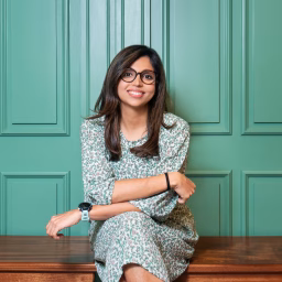 A woman with dark hair and glasses sits casually on a bench in front of a green wall