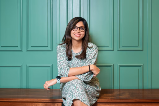 A woman with dark hair and glasses sits casually on a bench in front of a green wall