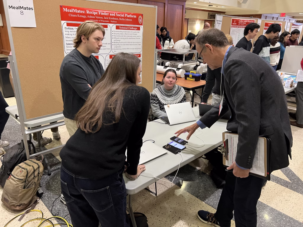 Alumni judge visits table of team demonstrating mobile app
