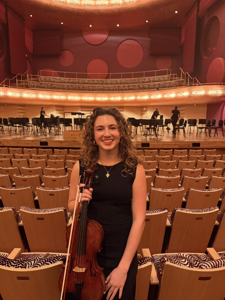 Janna Postma holding her viola in front of an orchestra stage.