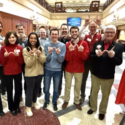 9 adults of varying ages standing in a group indoors holding up a "w" sign with their hands