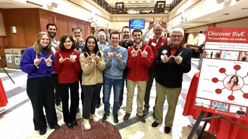 9 adults of varying ages standing in a group indoors holding up a "w" sign with their hands