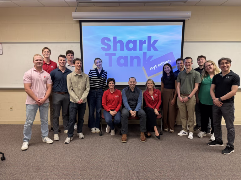A group of college students surround three seated adult professionals with a graphic behind them