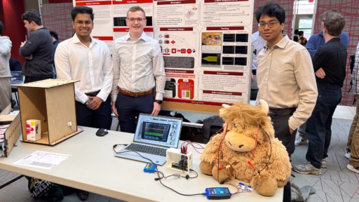 Three students standing in front of research poster and behind table with computer equipment and test stuffed animal