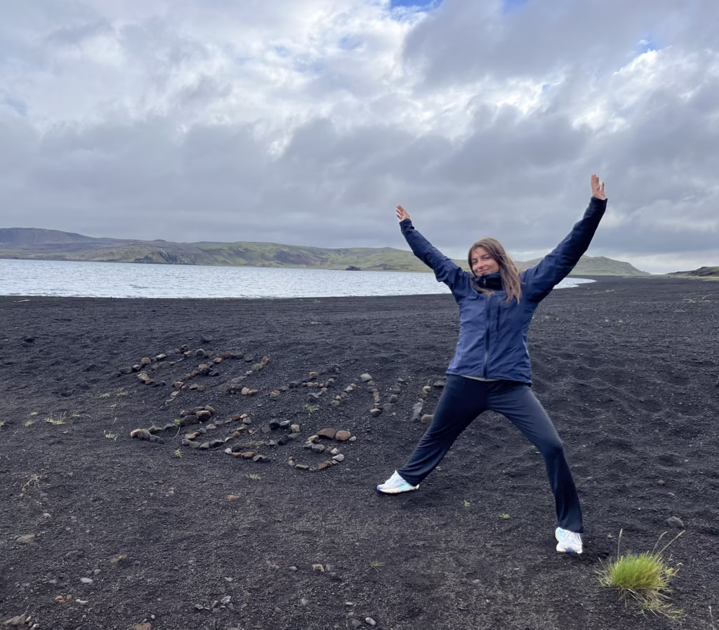 Anna Rosu is hiking on a beach and posing in front of rocks placed to spell "Iceland 2025."