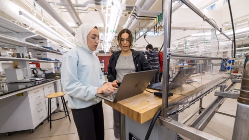 Two students stand in front of a computer in a lab working on a report.