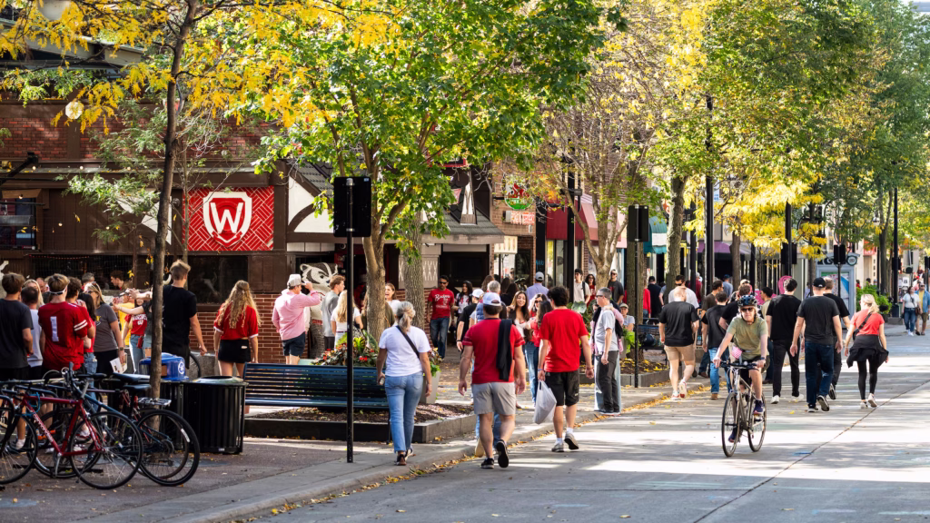 Pedestrians walk and bicyclists pedal up and down the 600 block of State Street in downtown Madison, Wis., during an autumn (fall) football game-day on Oct. 18, 2025. At left with a W emblem is State Street Brats, a popular restaurant and sports bars located near the University of Wisconsin–Madison campus.