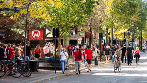 Pedestrians walk and bicyclists pedal up and down the 600 block of State Street in downtown Madison, Wis., during an autumn (fall) football game-day on Oct. 18, 2025. At left with a W emblem is State Street Brats, a popular restaurant and sports bars located near the University of Wisconsin–Madison campus.
