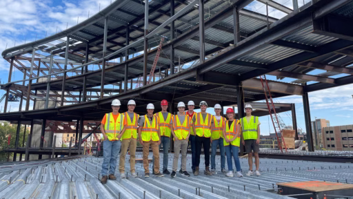 Students wearing safety vests and hard hats at the site of a building under construction.